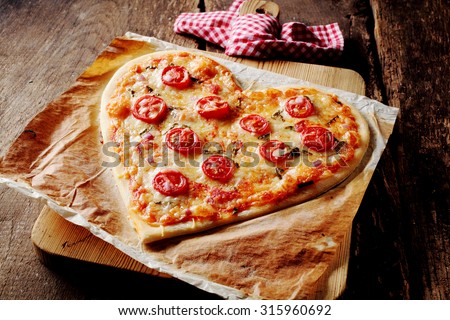 Baked heart-shaped homemade pizza topped with mozzarella and tomato slices, on parchment paper on a cutting board near a checkered red and white kitchen towel, on a rustic table, high-angle close-up