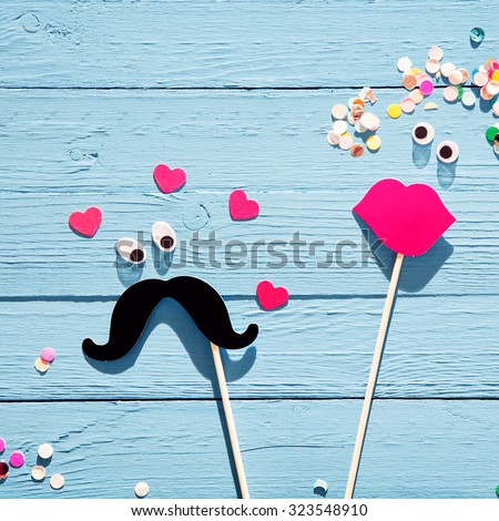 Fun romantic couple from photo booth accessories with a mustache with eyes surrounded by hearts eyeing a lady with luscious red lips and confetti flowers in her hair, on a rustic blue wood background