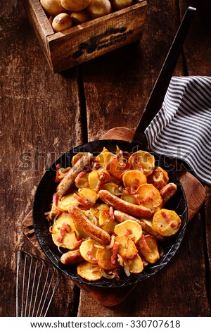 Tasty German recipe of golden roasted or fried potato slices, spicy sausage and bacon served on a rustic wooden counter in a vintage frying pan with a box of fresh potatoes alongside