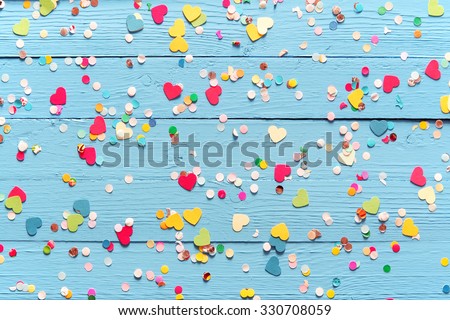 Blue wood background with scattered colorful party confetti with heart shapes in a closeup full frame overhead view for festive or celebration themed concepts