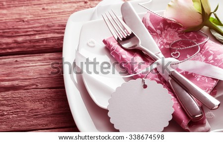 Blank Tag Tied Around Silver Knife and Fork Resting on Pink Napkin on top of White Dishes and Decorated with White Rose and Small Pearl Hearts - Romantic Wedding Place Setting with Copy Space