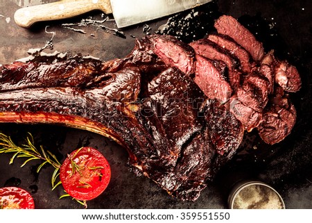 Sliced rare grilled or barbecued tomahawk beef steak on a griddle with fresh rosemary and tomato in a close up view