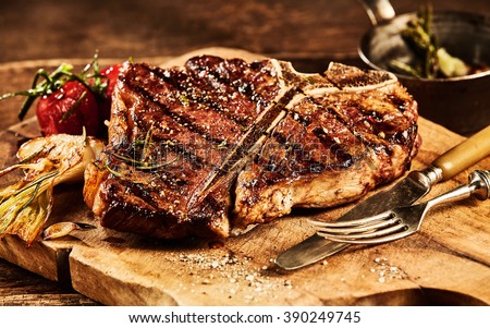 Succulent grilled large t-bone steak garnished with herbs, tomato and salt with fork and knife beside it on cutting board