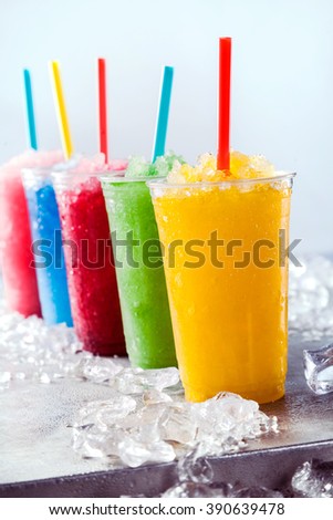 Still Life of Colorful Frozen Fruit Granita Slush Drinks in Plastic Take Away Cups with Drinking Straws Arranged in a Row on Cold Metal Surface Surrounded by Ice Cubes