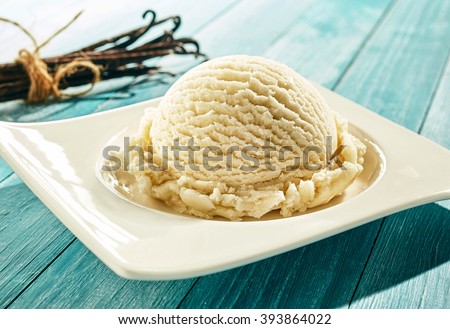 Single scoop of creamy vanilla ice cream on a stylish modern plate on a blue wooden picnic table outdoors in summer with a bundle of dried vanilla pods behind