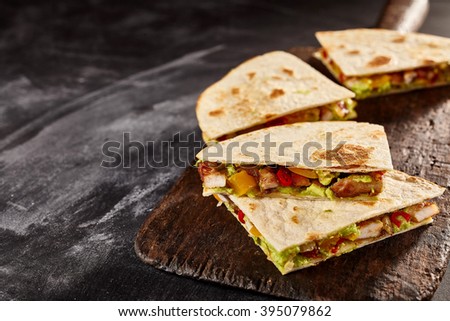 Four sliced wedges of meat and veggie filled quesadillas on worn out black cutting board over dark background with copy space