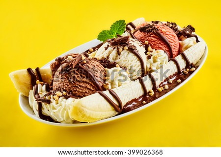 Tropical banana split with chocolate drizzle over three scoops of chocolate, strawberry and vanilla ice cream on fresh bananas, yellow background