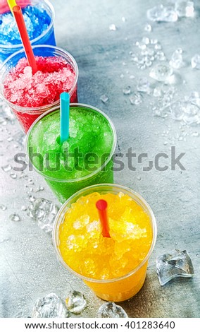 High Angle Still Life View of Colorful Frozen Fruit Granita Slush Drinks in Plastic Cups with Drinking Straws Arranged in a Row on Cold Metal Counter with Scattered Ice Cubes with Copy Space