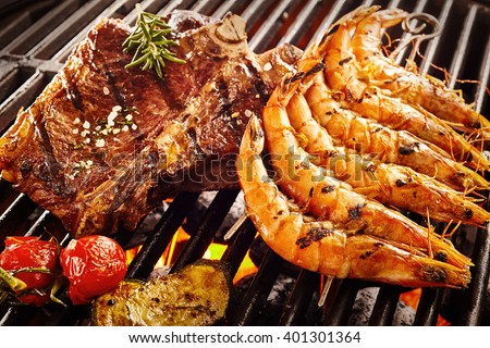 Side of beef garnished with rosemary and salt next to prawns and tomatoes roasting on flaming grill
