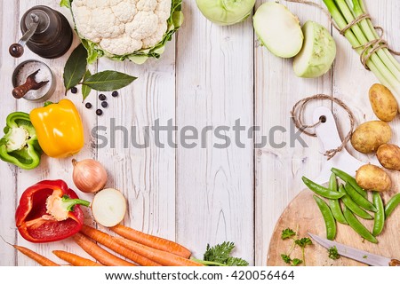 Delicious fresh vegetables forming a frame on white wood with central copy space together with a cutting board, knife and seasoning for cooking