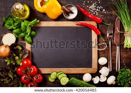 High Angle View of Blank Chalkboard and Silver Knife and Fork Surrounded by Fresh Herbs and Healthy Raw Vegetables Scattered on Rustic Wooden Table Surface