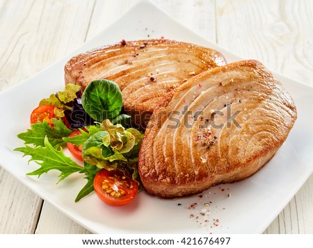 Close Up Still Life of Two Fried Tuna Steaks on Square White Plate with Seasoning and Fresh Green Salad with Tomato on Painted Wooden Table
