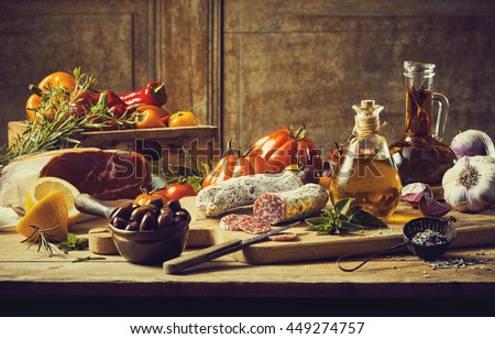 Retro style kitchen still life with fresh vegetable ingredients, cuts of cold meat and salami, condiments and olive oil on a rustic wooden table