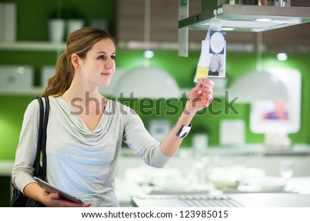 Young Woman Shopping For Furniture In A Furniture Store, Using Her Tablet Computer To Compare Prices/Check For Dimensions