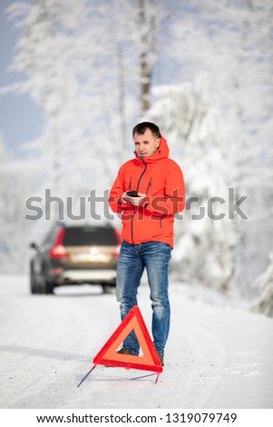 Handsome man setting up a warning triangle and calling for assistance after his car broke down in the middle of nowhere on a freezing winter day