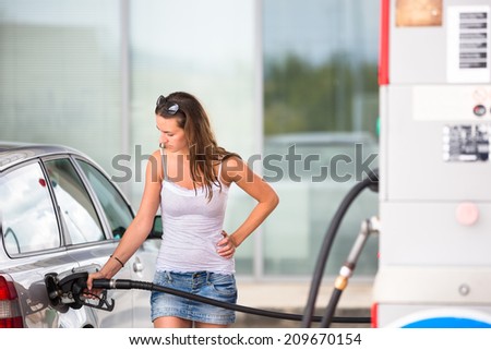 Attractive, young woman refueling her car in a gas station, checking the amount of gas, disliking the price tag and the gas mileage of her engine