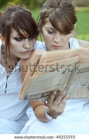 two pretty young women reading together a newspaper outdoors on a lovely summer day