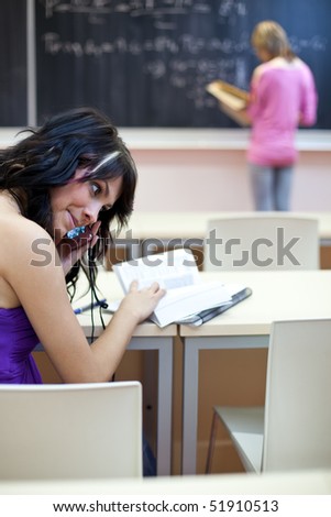 pretty female college student in a classroom using her cellphone while the teacher writes on the blackboard