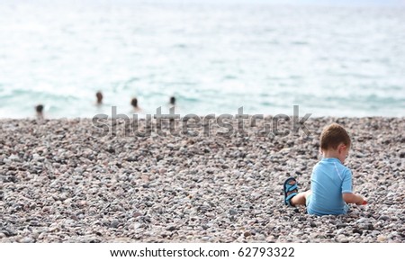 little boy playing on the beach alone while members of his family swim in the sea