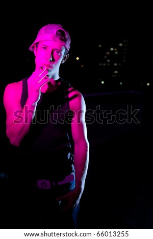 handsome young man lit with colorful lights standing on a roof at night, smoking a cigarette  (color toned image)