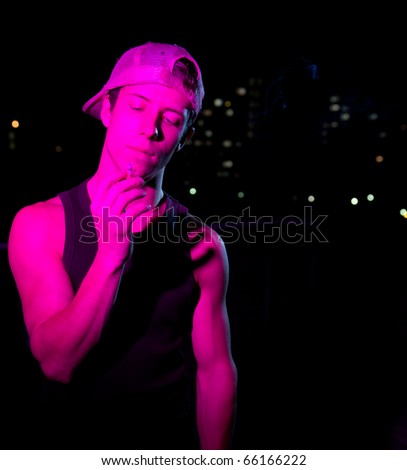 handsome young man lit with colorful lights standing on a roof at night, smoking a cigarette (color toned image)
