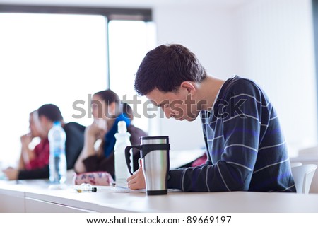 young, handsome male college student sitting in a classroom full of students during class (color toned image; shallow DOF)