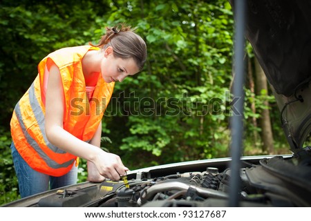 Young female driver wearing a high visibility vest, bending over the engine of her broken down car