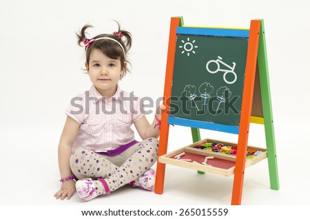Amazed baby girl draw flowers, a bike and Sun on black board with chalk isolated on white