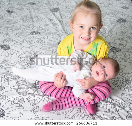 A girl holding in her arms her younger three month old sister sitting on a bed.