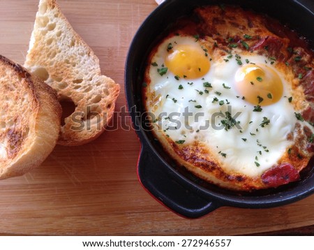 Shakshuka breakfast with sourdough toast. Black clay pot on a wood board base