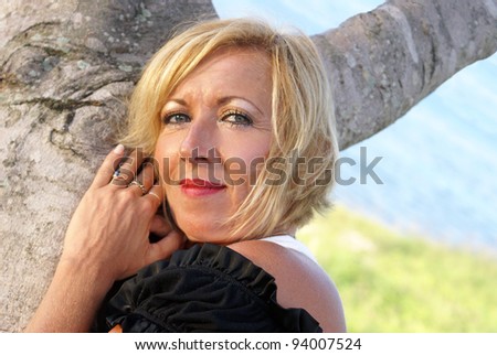 A woman is looking at the viewer while leaning on a tree near the beach.