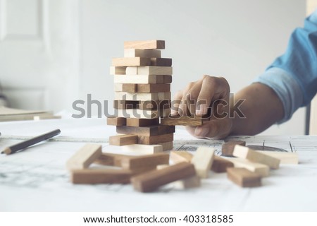 Hand of engineer playing a blocks wood game (jenga) on blueprint or architectural project vintage tone.