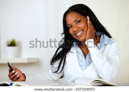 Portrait of a friendly young woman looking to you while holding a cellphone in one hand at home indoor