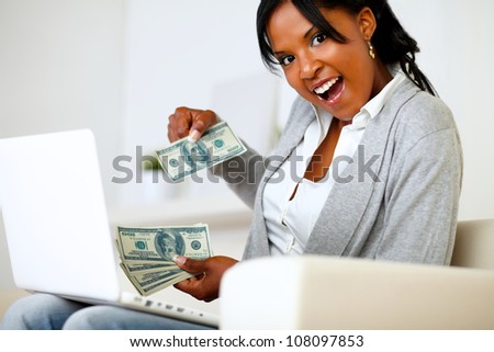 Portrait of a surprised woman with dollars is looking to you while is sitting on sofa at home in front a laptop