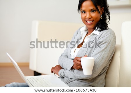 Portrait of a young woman with a mug is looking to you and browsing the Internet on her laptop while is sitting on the floor at home indoor