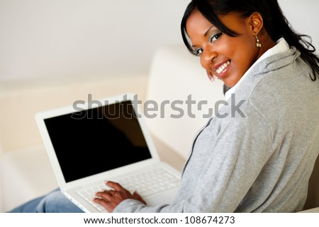 Portrait of a relaxed afro-American female browse the Internet on laptop while is sitting on sofa at home indoor and is looking to you