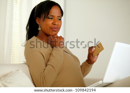 Portrait of a pretty woman thinking whether to buy online with her credit card while sitting on sofa in front her laptop at home indoor
