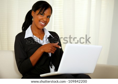 Portrait of a woman on black suit pointing to laptop screen while sitting on sofa at home indoor and looking at you