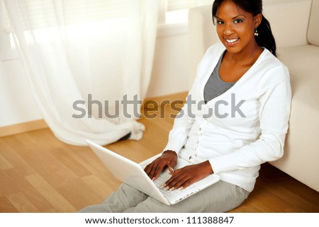 Top view portrait of a young woman looking at you and browsing the Internet while sitting on the floor at home indoor. With copyspace