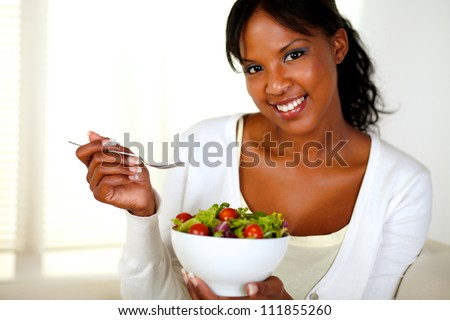 Portrait of a young woman looking at you while eating fresh salad at home indoor