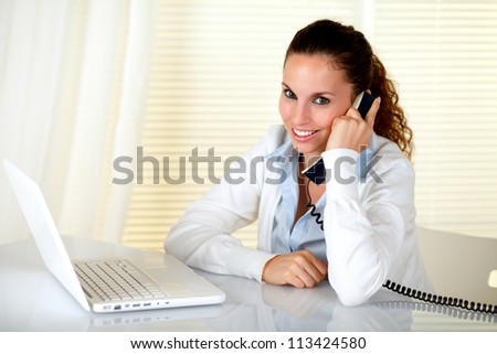 Lovely woman smiling and looking at you while speaking on phone in front of her laptop