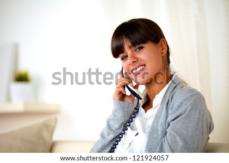 Portrait of a young woman conversing on phone looking at you while sitting on sofa at home indoor