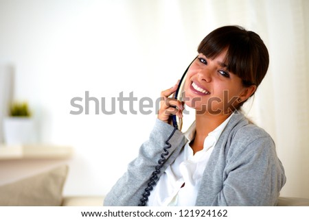 Portrait of a young female conversing on phone looking at you while sitting on sofa at home indoor