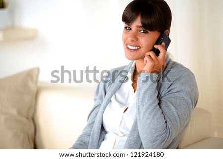 Portrait of a young woman looking at you conversing on cellphone while is sitting on sofa at home
