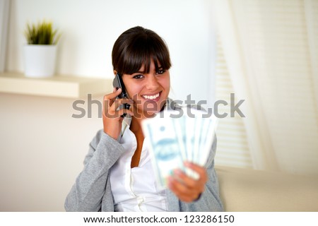 Portrait of a smiling young woman looking at you and speaking on cellphone with money