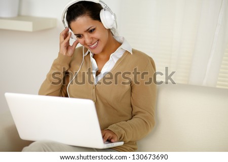 Portrait of a young female with headphone reading on laptop screen while is sitting on couch at home indoor