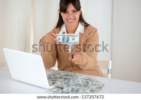 Portrait of a young woman holding and showing you cash dollars in front of her laptop while is pointing at you