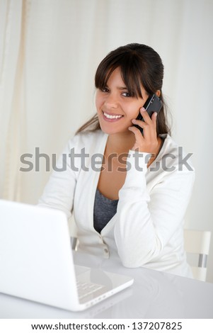 Portrait of a smiling young woman looking at you and using her laptop while is speaking on cellphone
