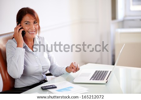Portrait of a beautiful professional woman conversing on her cell and smiling at you while sitting on office desk