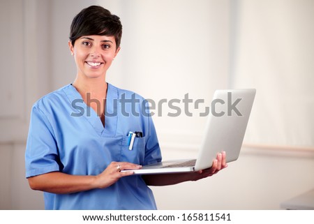 Portrait of a feamle latin doctor on blue uniform working on her laptop while standing and smiling at you on hospital